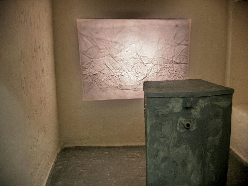 A glowing, backlit panel etched with branching vein-like lines hangs on a wall, paired in the same room with a solid concrete or stone block on the floor in front of it.