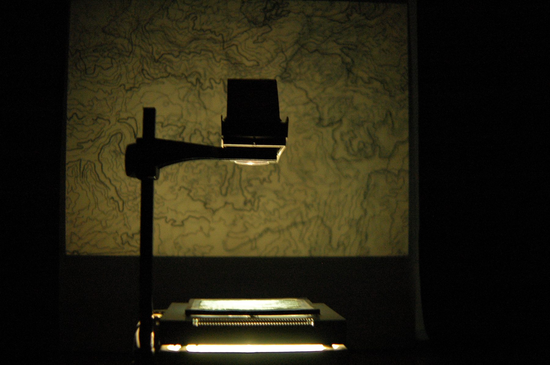 An overhead projector casting a hand-drawn topographic map onto a wall at the end of a dark corridor, the projector lamp glowing brightly in the foreground.