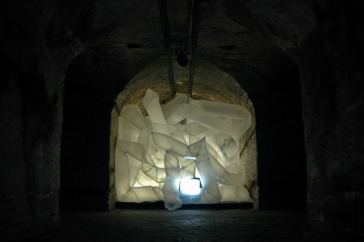 A dense cluster of translucent white inflated fabric tubes filling a vaulted brick alcove, internally lit and glowing brightly through a gap at the base.